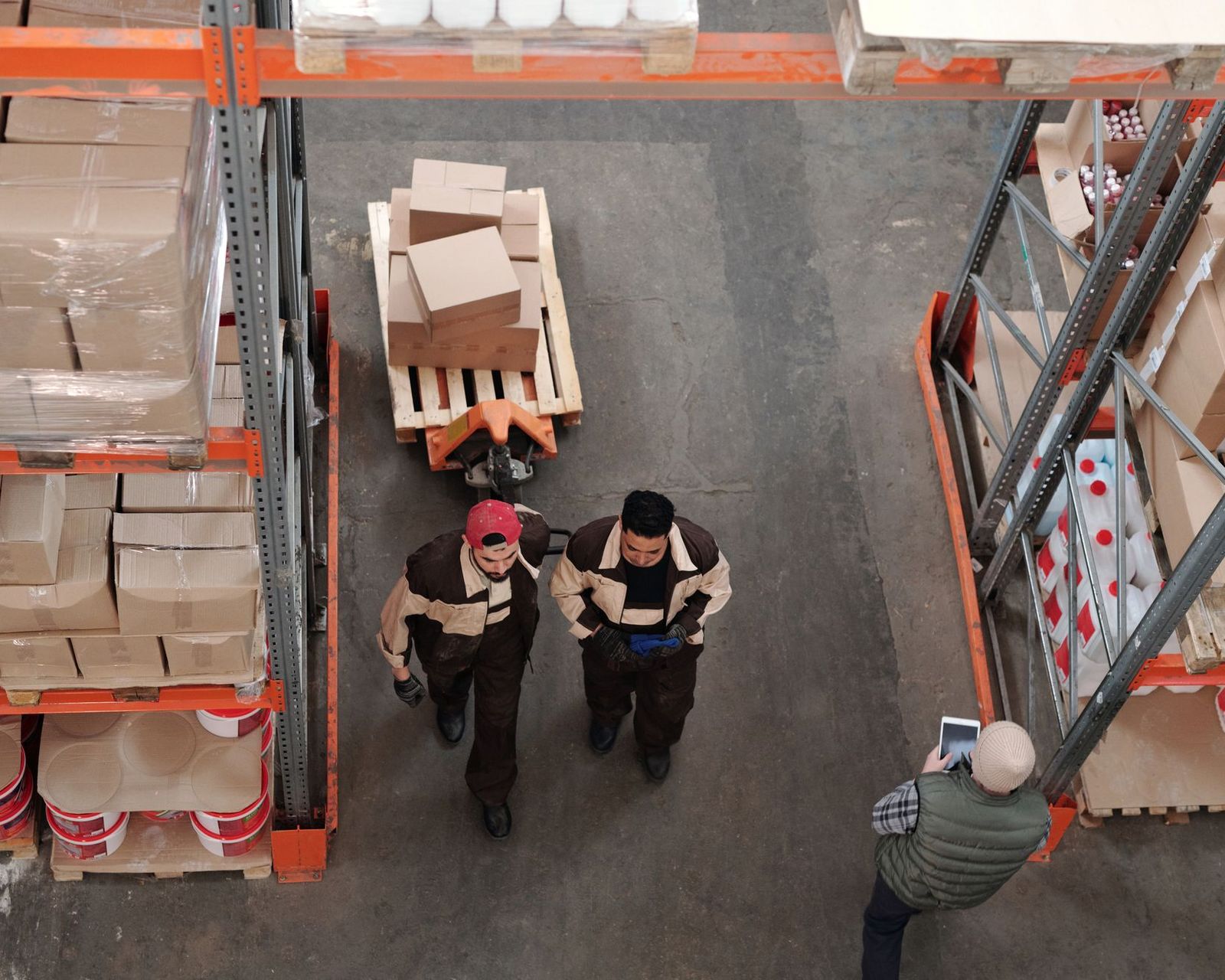 Overhead view of two warehouse workers walking between pallet racking of boxed stock while a manager checks figures on a tablet